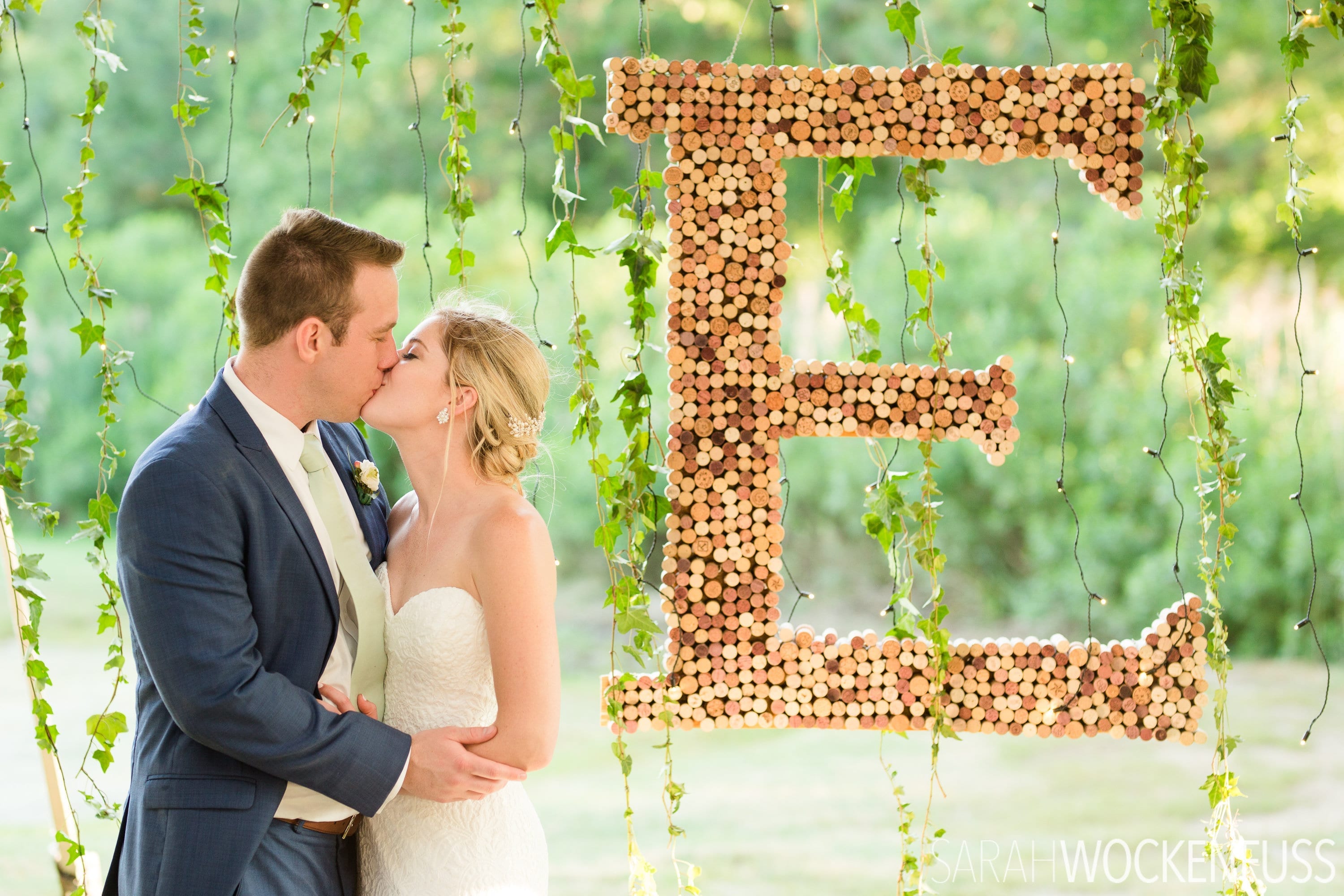 Wedding couple kissing near large letter E made of wine corks.