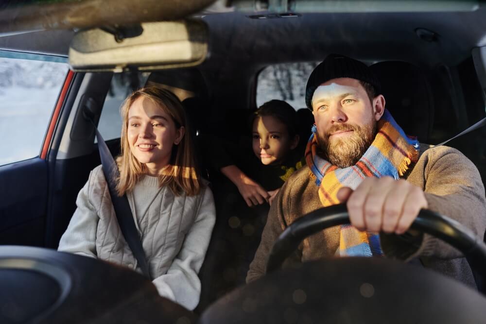 A father and mother dressed warmly in the front seat of a car with their daughter in the backseat during a winter road trip